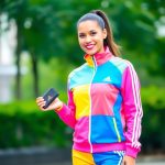A confident woman in a colorful Adidas tracksuit (pink, yellow, and blue) stands outdoors, holding a small Adidas wallet, promoting Adidas affiliate programs commission with a smile.