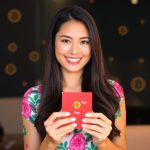 A smiling woman with long dark hair holding a red Make money online with Binance Pay Red Packet giveaways, wearing a vibrant floral dress, with digital currency symbols in the background.
